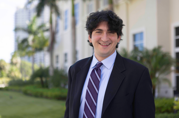 A picture of Ari Sasson, smiling wearing a suit and a purple striped tie.He is outside with palm trees in the background